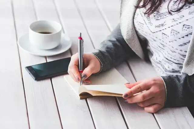 A woman sits at a desk, writing in her journal with a cup of coffee nearby.
