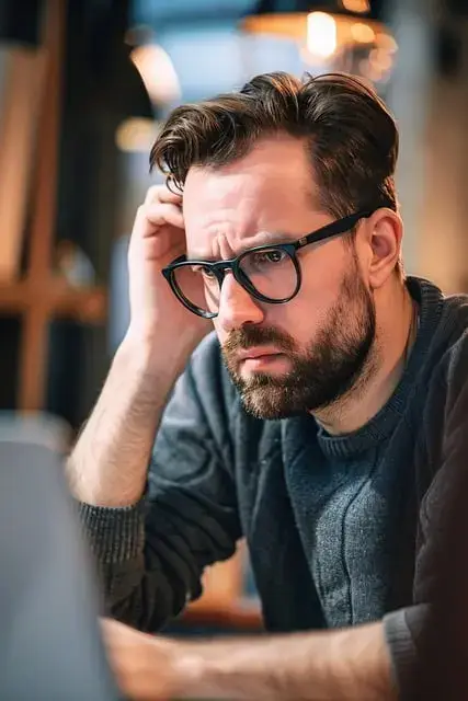 A man sits at a computer, scratching his head in confusion