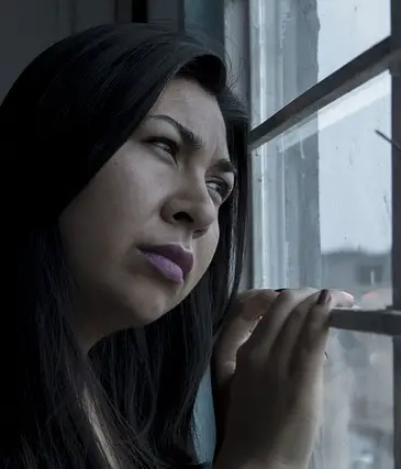 A Hispanic woman looks out a window. Her expression is one of sadness and stress.