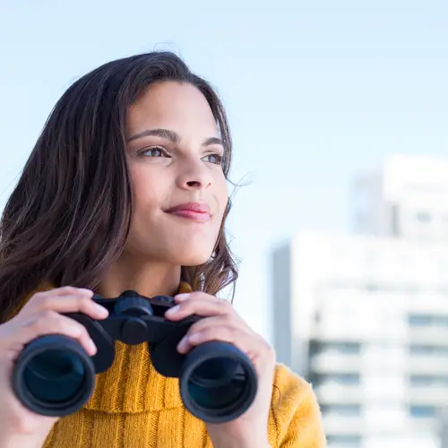 A woman looks into the distance with binoculars poised to see through.