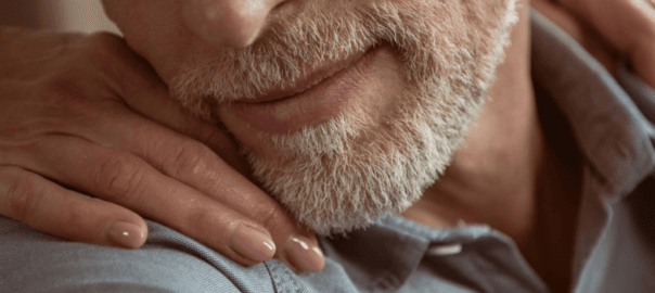 A close up photo of an older man's chin as he turns to embrace the younger hands which are laying on his shoulders.